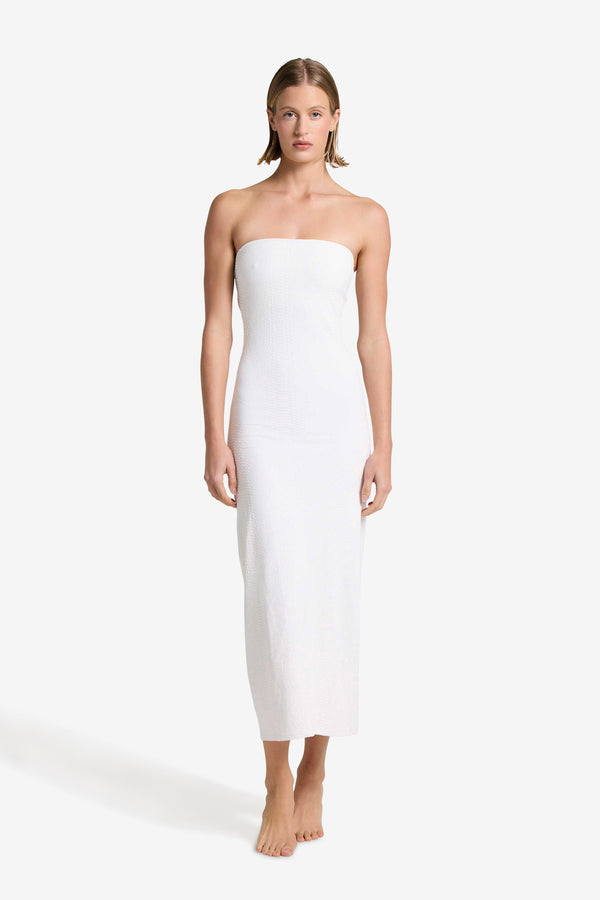 Barefoot against a plain backdrop, a person wears Gigi C Bikinis elegant Maya Dress in ivory. The strapless, ankle-length design complements their relaxed posture as short, straight hair frames their serene demeanor, with arms at their sides.