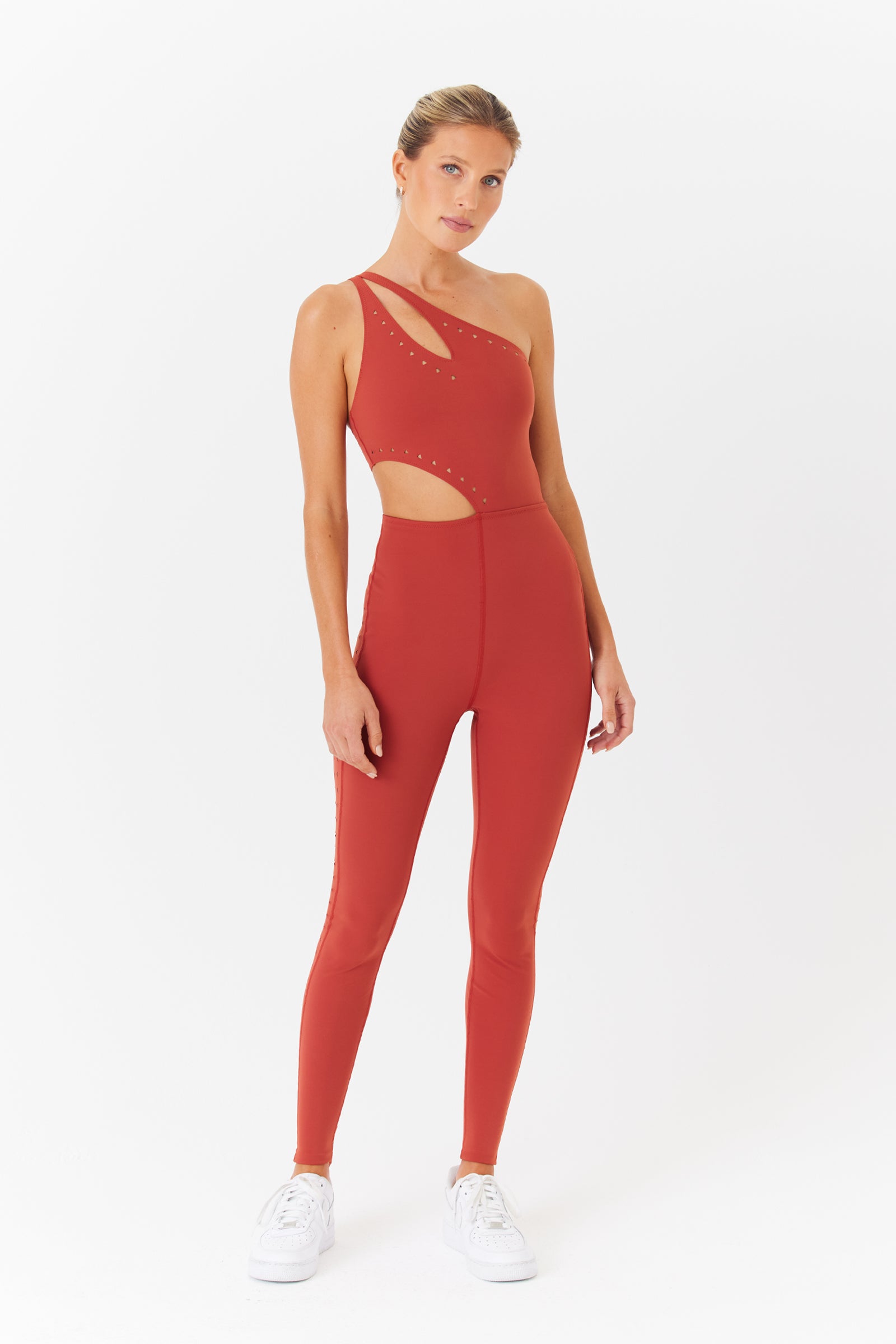 A woman in a red Cassie jumpsuit with asymmetrical design, laser cutouts, and embellishments by Gigi C Sport stands on a white background. The breathable knit fabric pairs stylishly with her tied-back hair and white sneakers.