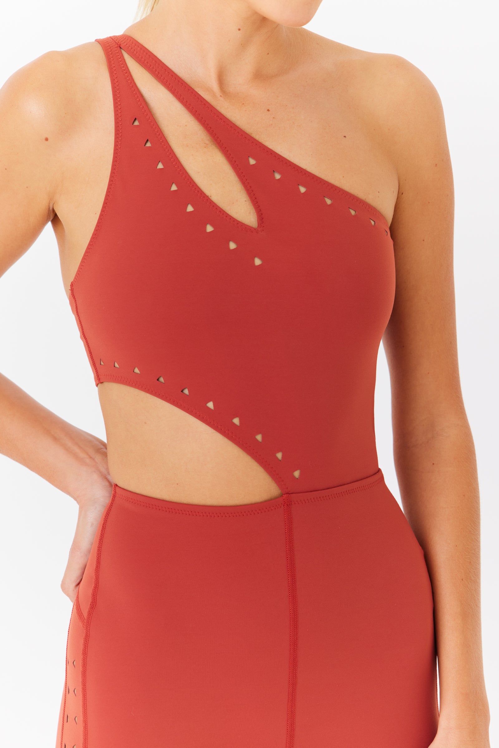 A person in a red Cassie Jumpsuit by Gigi C Sport features an asymmetrical one-shoulder design with laser cutouts and small triangular embellishments, highlighted from chest to waist, with the persons hand resting confidently on their hip.
